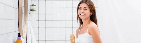 Glückliche junge Frau lächelt in die Kamera, während sie eine Haarbürste im Badezimmer hält, Banner — Stockfoto