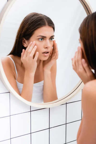 Worried young woman looking in mirror and touching face in bathroom, blurred foreground — Stock Photo