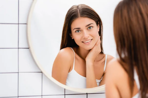 Heureuse jeune femme touchant le visage et souriant près du miroir dans la salle de bain, flou au premier plan — Photo de stock