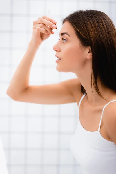 Vista lateral da jovem mulher em singlet pinça sobrancelhas no banheiro — Fotografia de Stock