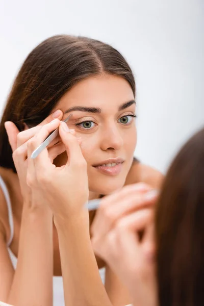 Young woman tweezing eyebrows near mirror, blurred foreground — Stock Photo