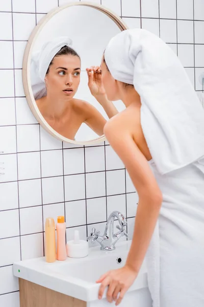 Молодая женщина с белым полотенцем на голове выщипывает брови в ванной комнате — стоковое фото