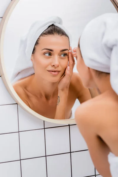 Giovane donna con asciugamano di spugna bianco sulla testa pinzetta sopracciglia in bagno vicino allo specchio, primo piano sfocato — Foto stock