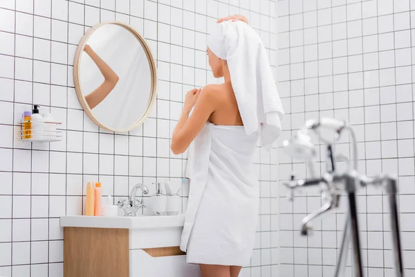 Mujer joven, envuelta en toallas de rizo blanco, de pie en el baño cerca del espejo en primer plano borroso - foto de stock