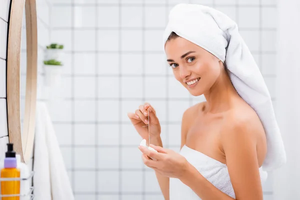 Femme souriante avec serviette blanche sur la tête souriant à la caméra tout en tenant fil dentaire dans la salle de bain — Photo de stock