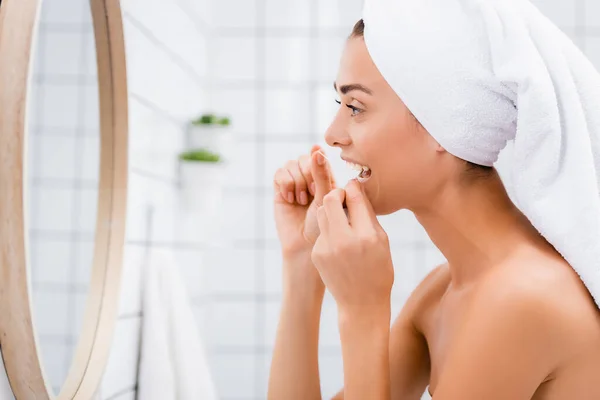 Vista laterale della giovane donna con asciugamano bianco sulla testa denti filo interdentale vicino specchio in bagno — Foto stock