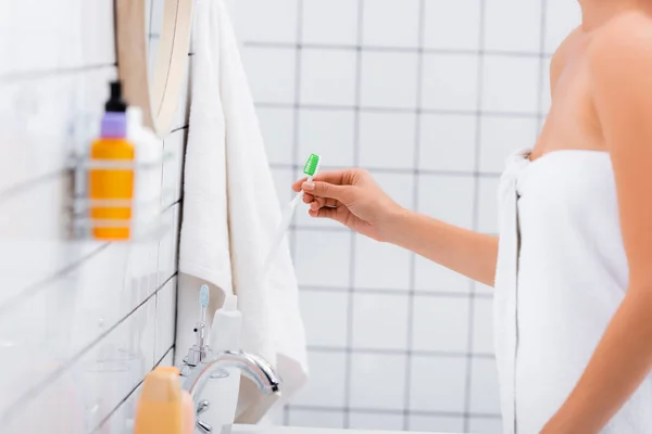 Обрезанный вид женщины, завернутый в белое полотенце, держащий зубную щетку в ванной комнате, размытый передний план — стоковое фото