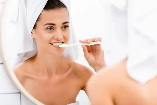 Young woman with white towel on head brushing teeth while looking in mirror, blurred foreground — Stock Photo