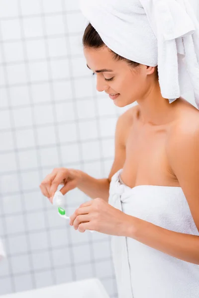 Молодая женщина, завернутая в белые полотенца, наносит зубную пасту на зубную щетку в ванной комнате — стоковое фото