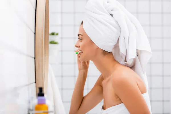 Vista laterale della donna con asciugamano bianco sulla testa lavarsi i denti in bagno, primo piano sfocato — Foto stock