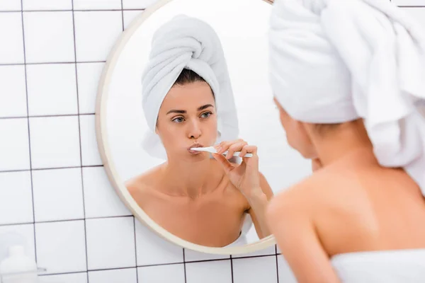 Mulher com toalha branca na cabeça olhando no espelho enquanto escova os dentes no banheiro, foreground borrado — Fotografia de Stock