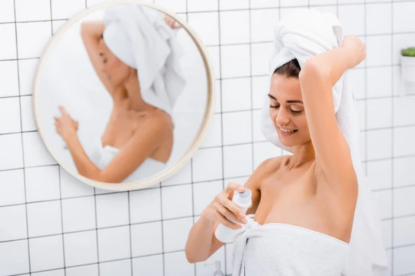 Счастливая женщина с белым махровым полотенцем на голове с помощью дезодоранта в ванной комнате рядом с размытым отражением в зеркале — стоковое фото