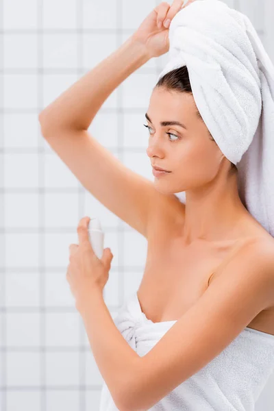 Mujer joven con toalla de rizo blanco en la cabeza rociando desodorante en el baño - foto de stock