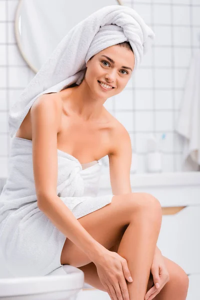 Cheerful woman with white towel on head smiling at camera while applying body lotion on leg in bathroom — Stock Photo