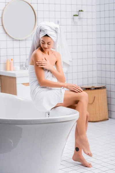 Mujer joven, envuelta en toallas de rizo blanco, aplicando loción corporal mientras está sentada en la bañera - foto de stock