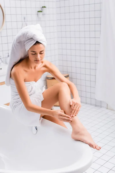 Mujer joven con toalla blanca en la cabeza aplicando loción corporal en la pierna mientras está sentada en la bañera — Stock Photo