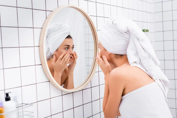 Joven mujer disgustada con toalla de rizo blanco en la cabeza tocando la cara mientras se mira en el espejo - foto de stock