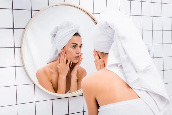 Dissatisfied young woman with white towel on head looking in mirror and touching face in bathroom — Stock Photo