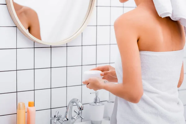 Частичный вид женщины, завернутой в белое полотенце, держащей косметический крем в ванной комнате — стоковое фото