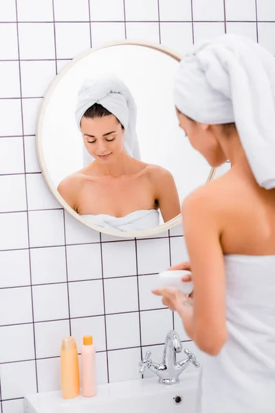 Молодая женщина, завернутая в белые полотенца, держащая косметический крем в ванной комнате, размытый передний план — стоковое фото