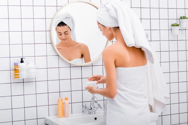 Giovane donna con asciugamano bianco sulla testa apertura crema cosmetica in bagno — Foto stock