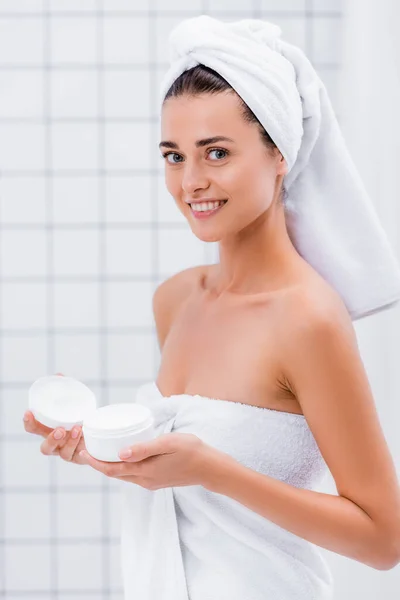 Joyeuse jeune femme souriant à la caméra tout en tenant la crème cosmétique dans la salle de bain — Photo de stock
