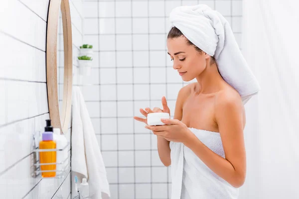 Молодая женщина с белым полотенцем на голове трогает косметический крем в ванной комнате — стоковое фото