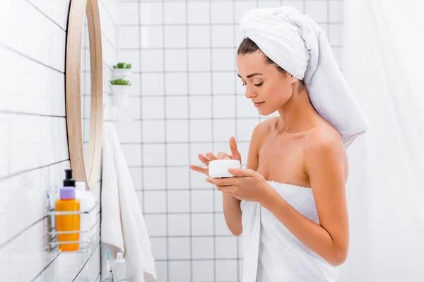 Jovem com toalha branca na cabeça tocando creme cosmético no banheiro — Fotografia de Stock