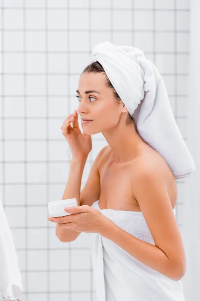 Mujer joven con toalla en la cabeza aplicando crema facial en el baño - foto de stock