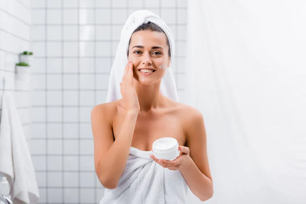 Щаслива молода жінка, загорнута в рушники, наносить крем для обличчя і посміхається на камеру у ванній — стокове фото