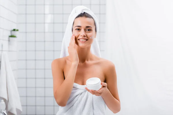 Jovem feliz, envolto em toalhas, aplicando creme facial e sorrindo para a câmera no banheiro — Fotografia de Stock