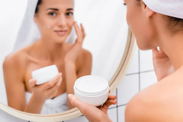 Young woman applying face cream near blurred mirror reflection in bathroom — Stock Photo