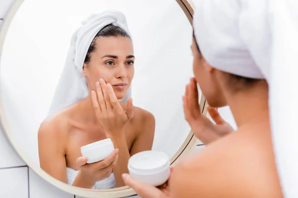 Mujer con toalla blanca en la cabeza mirando en el espejo mientras se aplica crema facial en el baño - foto de stock