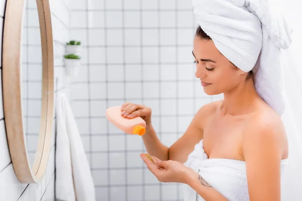 Улыбающаяся женщина с белым полотенцем на голове держа бутылку с лица скраб в ванной комнате — стоковое фото