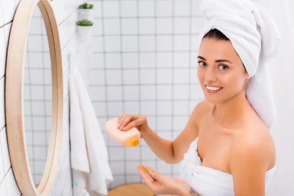 Femme souriante regardant la caméra tout en tenant bouteille avec gommage facial dans la salle de bain — Photo de stock