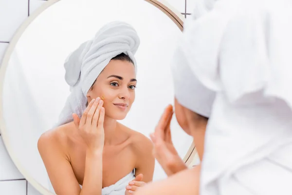 Jeune femme avec serviette blanche sur la tête appliquant gommage facial dans la salle de bain, avant-plan flou — Photo de stock