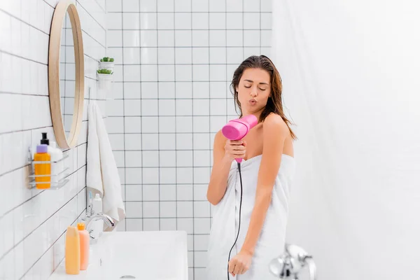 Молодая женщина, завернутая в белое полотенце, поет с феном в ванной комнате — стоковое фото