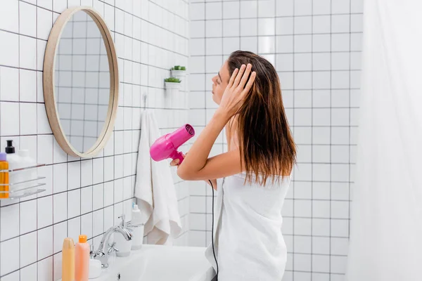 Jeune femme, enveloppée dans une serviette blanche, sèche-cheveux près du miroir dans la salle de bain — Photo de stock