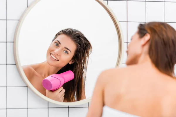 Junge Frau lächelt beim Haartrocknen in der Nähe von Spiegel im Badezimmer, verschwommener Vordergrund — Stockfoto