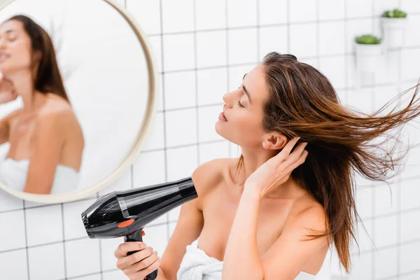 Young woman with closed eyes drying hair in bathroom near blurred reflection in mirror — Stock Photo
