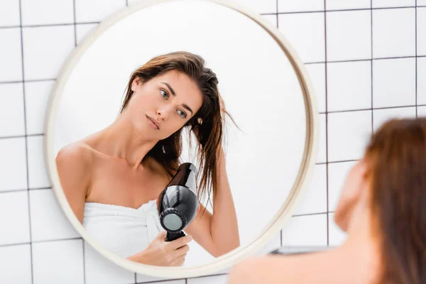 Jeune femme regardant dans le miroir tout en séchant les cheveux dans la salle de bain sur le premier plan flou — Photo de stock