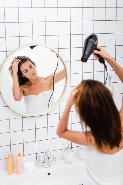 Jeune femme, enveloppé dans une serviette blanche, regarder dans le miroir tout en séchant les cheveux dans la salle de bain — Photo de stock