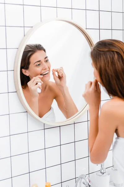 Молодая женщина зубной нитью рядом с зеркалом в ванной комнате, размытый передний план — стоковое фото