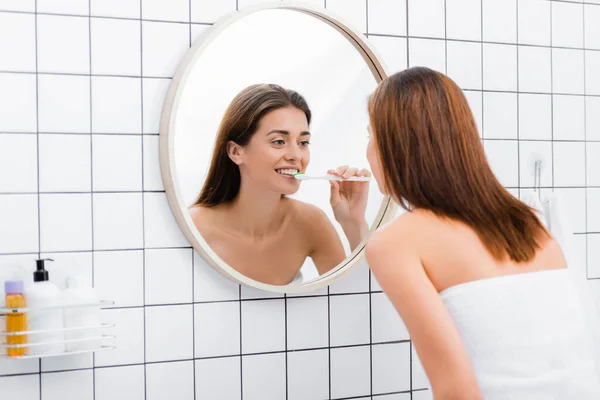 Mujer joven mirando en el espejo mientras se cepilla los dientes en el baño - foto de stock