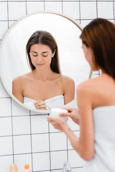 Молодая женщина, завернутая в белое полотенце, открывающая косметический крем возле зеркала, размытый передний план — стоковое фото