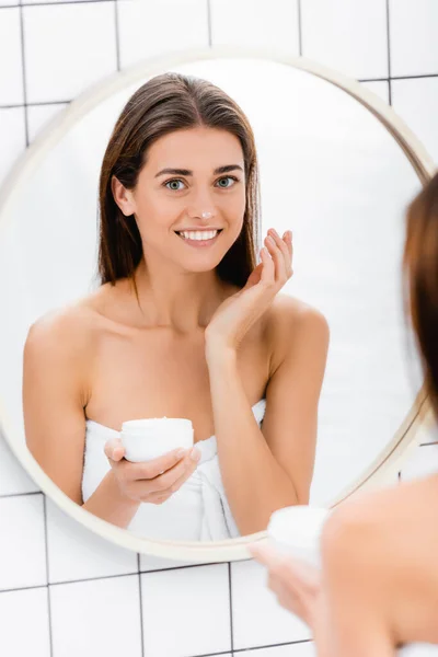 Mulher feliz sorrindo ao aplicar creme facial perto do espelho no banheiro, foreground borrado — Fotografia de Stock