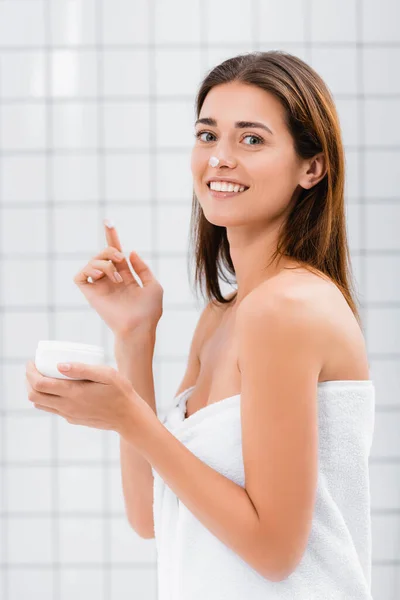 Feliz joven con crema facial en la nariz mirando a la cámara en el baño - foto de stock