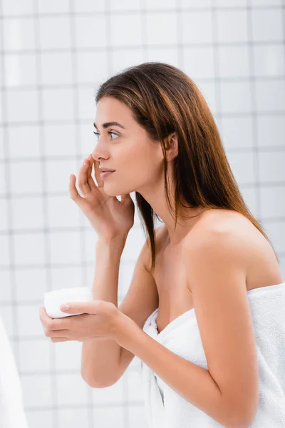 Молодая женщина, завернутая в белое полотенце, наносит крем для лица в ванной комнате — стоковое фото