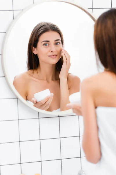 Femme heureuse appliquant crème visage près du miroir dans la salle de bain, avant-plan flou — Photo de stock