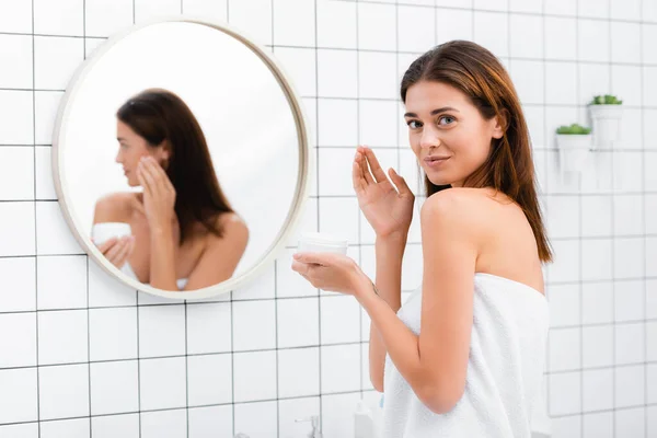 Happy young woman looking at camera while holding container of cosmetic cream in bathroom — Stock Photo